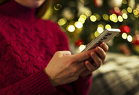 NDR-"Weihnachtstelefon" bietet Seelsorge und Gespräche Frau hält ein Smartphone, während sie in der Nähe eines mit Lichtern geschmückten Weihnachtsbaumes sitzt und einen roten Pullover trägt.