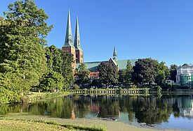 Auf dem Jakobsweg über Lübeck nach Hamburg: 125 Kilometer in fünf Tagen Der Lübecker Dom mit zwei Türmen im Morgenlicht, im Vordergrund eine Wasserfläche.
