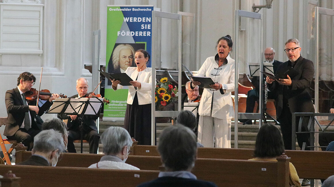 Geistliche Morgenmusik bei der Greifswalder Bachwoche Sängerinnen, Sänger und Musiker stehen im Dom zu Greifswald und musizieren. Aufgrund der Corona-Pandemie stehen Scheiben aus Plexiglas zwischen den Singenden.