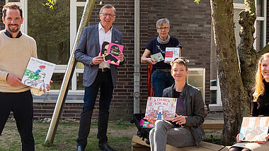 Fünf Frauen und Männer stehen und sitzen mit Abstand zueinander vor einer Hauswand. Sie halten Bücher für Kinder vor sich. 