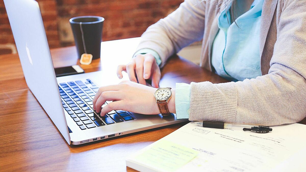 Person sitzt am Laptop, daneben ein aufgeschlagenes Buch