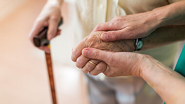 Zwei Hände einer Pflegerin halten die Hand einer älteren Patientin. In der anderen Hand hält sie den Griff eines Gehstocks.