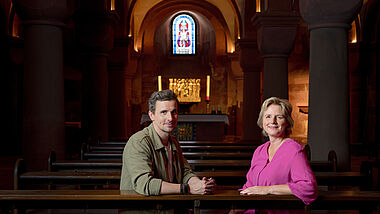 "die letzte Bank – Fragen an das Leben": Julian Sengelmann und Melanie Wolfers sitzen nebeneinander in der letzten Kirchenbank und blicken in die Kamera. Hinter ihnen eröffnet sich der Blick in die Krypta des Mainzer Doms.