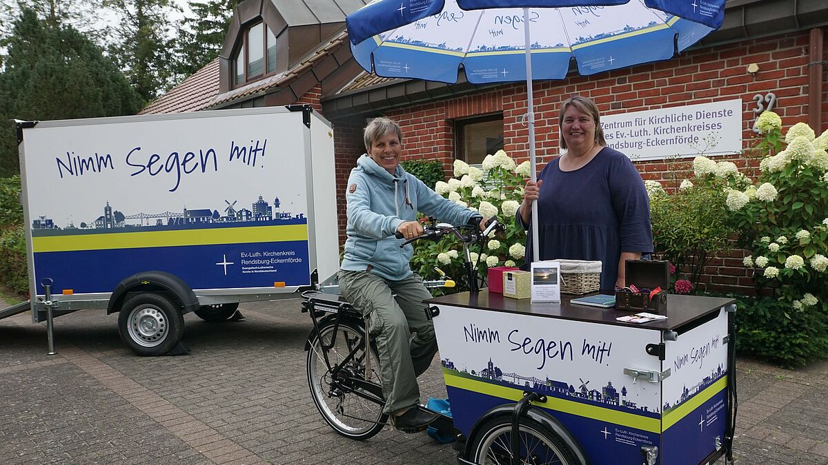 Tourismuspastorin Brigitte Gottuk auf dem Rad mit Diakonin Sabine Klüh. Daneben der PKW-Anhänger zur Beförderung des Segensrads. 