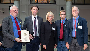 Fünf Personen, die Herren tragen Sakko und Krawatte, die Frau in der Mitte ein schwarzes Kostüm. Mann links hält den Förderbescheid in der Hand.