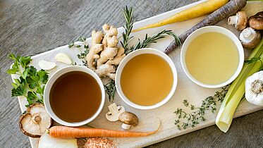 Auf dem Bild zu sehen sind drei kleine Schälchen mit Brühe in drei unterschiedlichen Farben. Drumherum verteilt liegen Zutaten für Brühe wie Zwiebel, Ingwer, Champignons. 