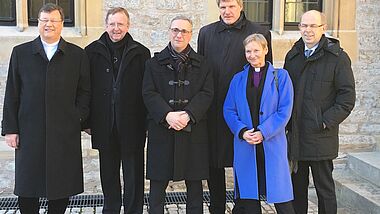 Bischöfe stehen alle in schwarz vor einem Gebäude, nur Bischöfin Fehrs trägt einen blauen Mantel