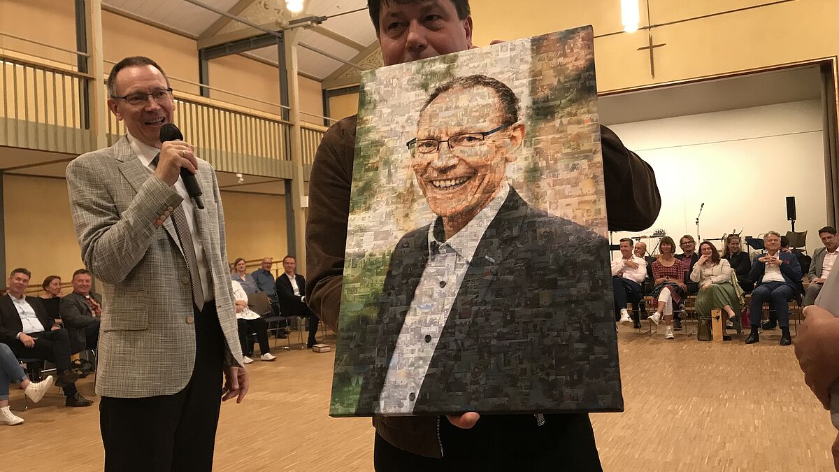 Wolfgang von Rechenberg erhält von der Evangelischen Schulstifung der Nordkirche ein Portraitfoto, zusammengesetzt aus ganz vielen kleinen Fotos aus dem Leben der evangelischen Schulen. Tim Bürger, Schulleiter des Greifswalder Seminars für kirchlichen Dienst SKD, hält es hoch.