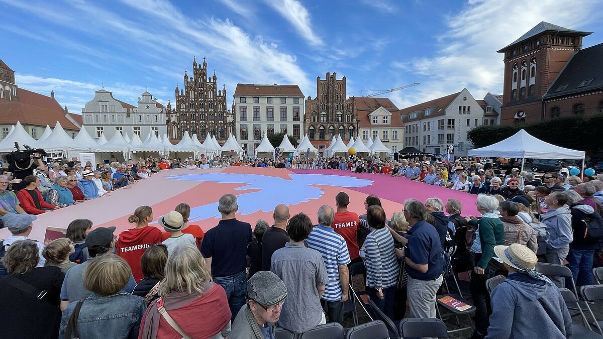 Auf dem Marktplatz breiten Bürger:innen und Kirchentagsteilnehmer:innen ein riesiges Friedenszeichen aus. 