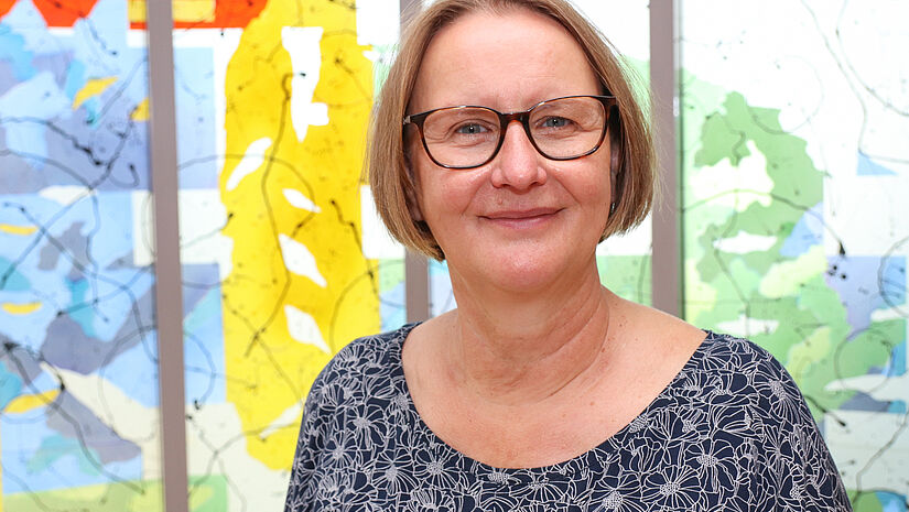 Porträtfoto einer Frau mit Brille, kinnlangen Haaren und bunter Bluse. Die steht vor modernen, farbigen Kirchenfenstern