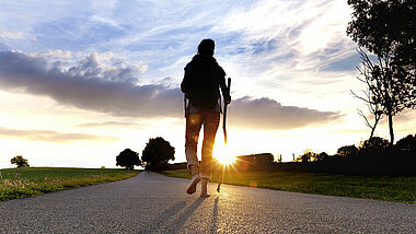 Eine Frau mit Wanderstock und Rucksack läuft barfuß auf einer Straße der untergehenden Sonne entgegen.