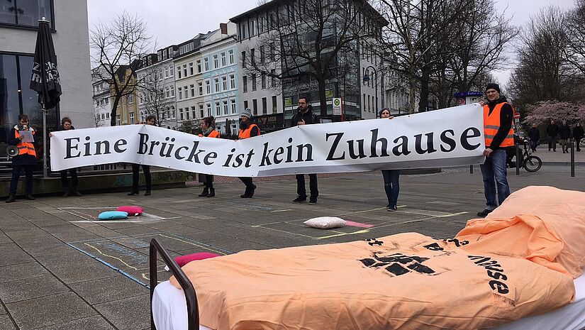 Menschen halten ein Banner in der Hand auf dem steht "Eine Brücke ist kein Zuhause", im Vordergrund steht ein Bett mit einer orangen Decke auf der Straße.