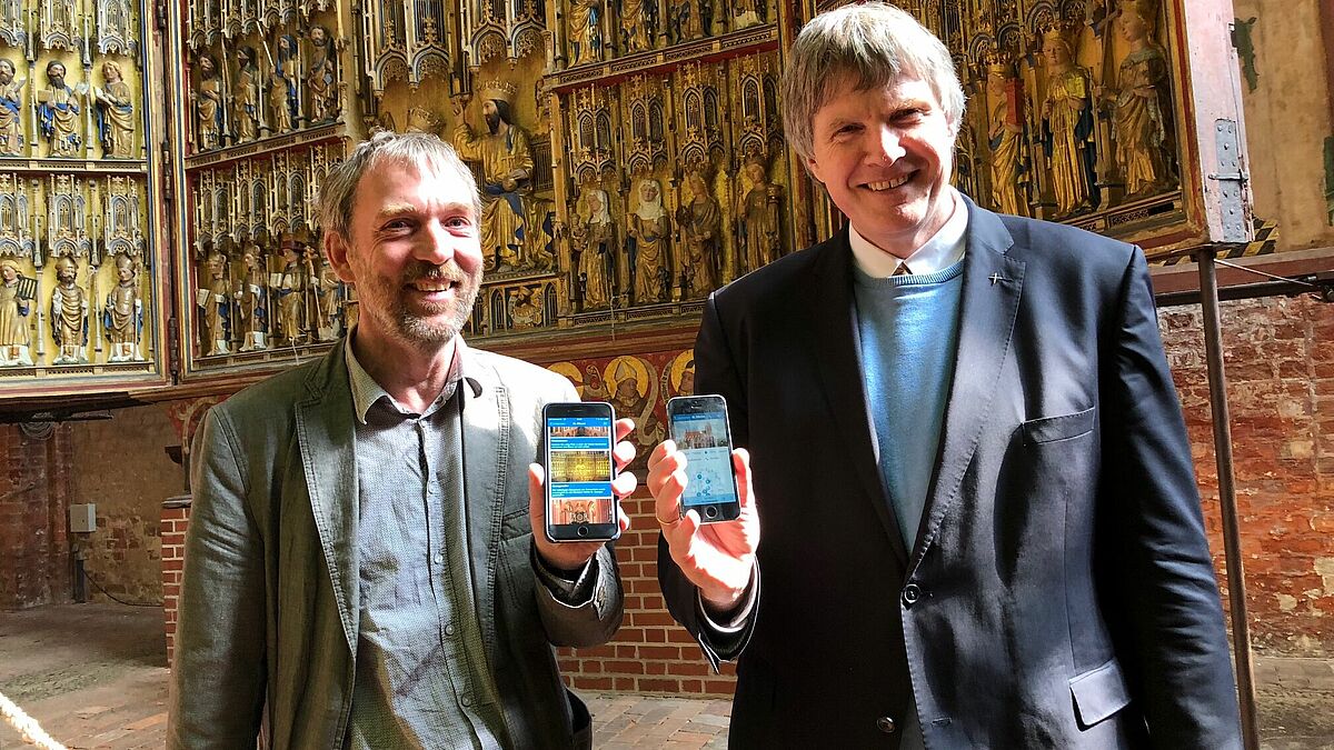 Gemeindepastor Roger Thomas (l.) und Bischof Andreas von Maltzahn zeigen die App "NordKirchenTour" vor dem Georgenaltar in der Kirche St. Nikolai in Wismar.