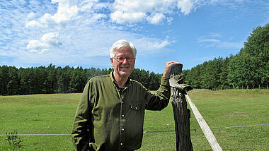 Heinrich Bedford-Strohm steht vor einer grünen Weide. Eine Hand lehnt auf einem Holzpfahl, im Hintergrund steht der hochsommerlich grüne Wald.