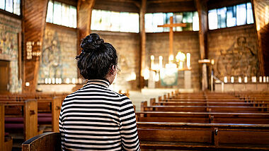 Das Bild zeigt Fofo, eine junge Frau aus dem Irak, in der Matthäuskirche in Darmstadt. Sie sitzt in der Kirche, ihre Haare sind zu einem Knoten hochgesteckt. Ihr Gesicht ist nicht zu erkennen.