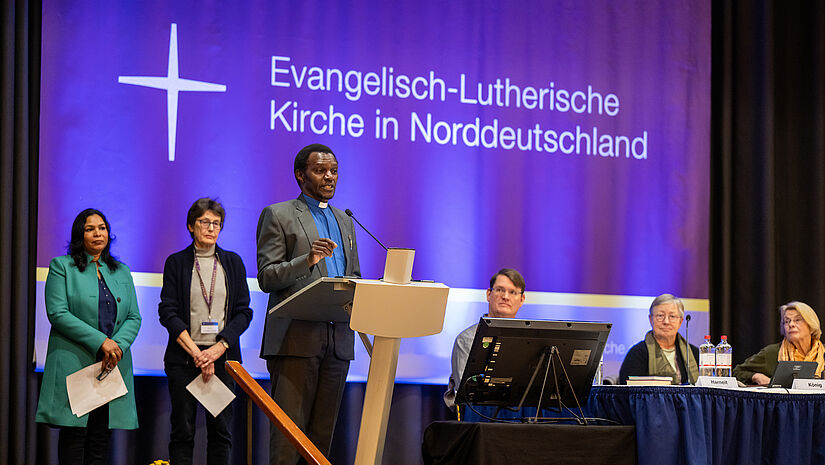 Ein Mann steht am Rednerpult, dahinter stehen und sitzen Menschen auf dem Podium.