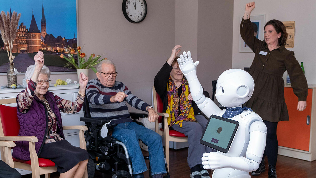 Jutta Tandler (rechts) von dem Pflegezentrum Travetal und Roboter Charlie machen mit Bewohnerinnen und Bewohnern Gymnastik.