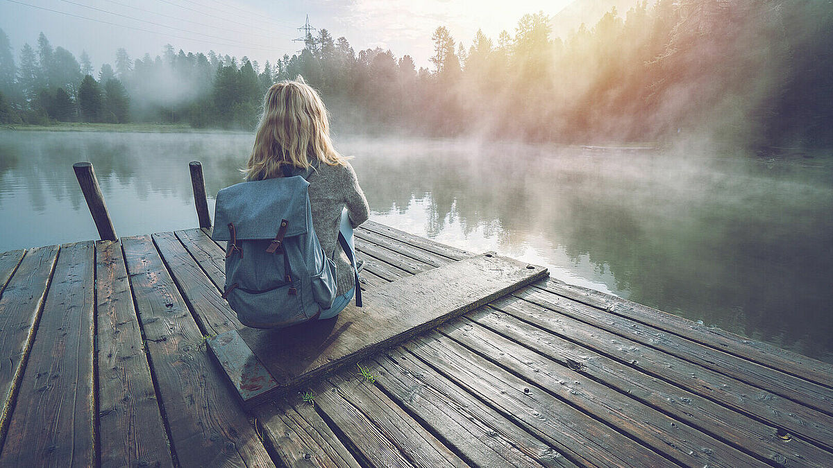 Frau sitzt mit dem Rücken zur Kamera aud einem Steg an einem See, der mit Tiefnebel bedeckt ist. Die Frau trägt einen Rucksack auf dem Rücken. Alles ist in Morgenlicht getaucht.