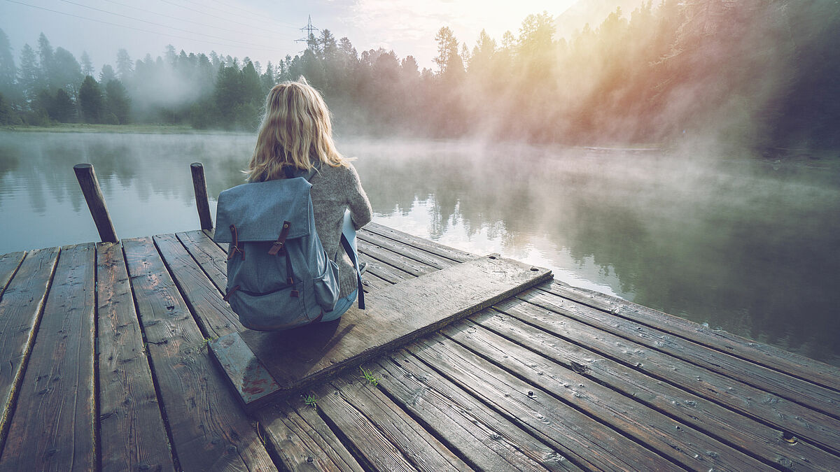 Frau sitzt mit dem Rücken zur Kamera aud einem Steg an einem See, der mit Tiefnebel bedeckt ist. Die Frau trägt einen Rucksack auf dem Rücken. Alles ist in Morgenlicht getaucht.