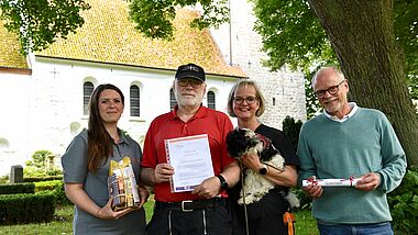 Küster Michael Hirner nimmt die Auszeichnung "ökofaire Gemeinde der Nordkirche" entgegen. Auf dem Foto zu sehen sind vor der Kirche St. Petri Bosau ebenso: Elena Stern vom Kirchenkreis (links), die stellvertretende Kirchengemeinderatsvorsitzende Sabine Hirner mit Hund Fine und Tobias Jahn vom Küsterarbeitskreis (rechts im Bild). 