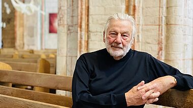 Stefan Schmidt sitzt in einer Kirchenbank. Er trägt einen dunklen, hoch geschlossenen Pullover und blickt direkt in die Kamera.