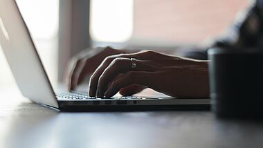 Hände tippen auf der Tastatur eines Laptops. Im Hintergrund bricht Licht durch ein Fenster in den Raum.