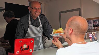 Der Bischof reicht einem Mann strahlend ein Fischbrötchen aus der Bude heraus