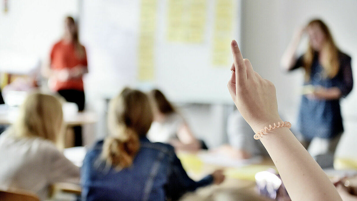 Im Vordergrund: Eine Hand, die sich meldet, im Hintergrund unscharf: Schüler, die an Tischen sitzen, eine Tafel, davor zwei Lehrpersonen