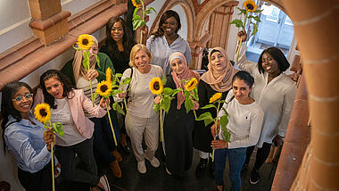 Frauen multiethnischer Herkunft stehen in einem Treppenhaus und halten strahlend Sonnenblumen in die Höhe.