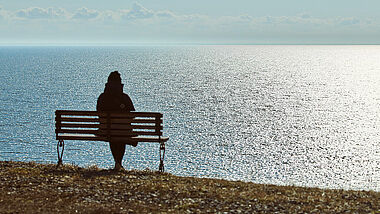 Eine Frau sitzt alleine auf einer Bank an einer Steilküste. Von ihr ist nur eine dunkle Silhouette zu sehen, das Meer glitzert in der Sonne.