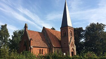 Der Auftakt der Reihe „Dorfkirche Mon Amour“ findet am 26. Mai rund um die Kirche im pommerschen Lüssow statt. Die Dorfkirche aus rotem Backstein in Lüssow steht umgeben von grünen Bäumen unter blauem Himmel.