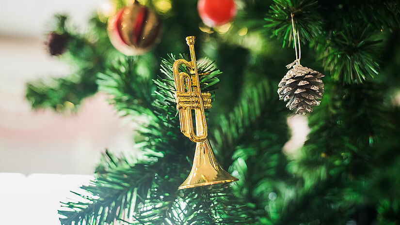 Eine kleine goldene Trompete hängt als Weihnachtsanhänger in einem festlich geschmückten Tannenbaum.