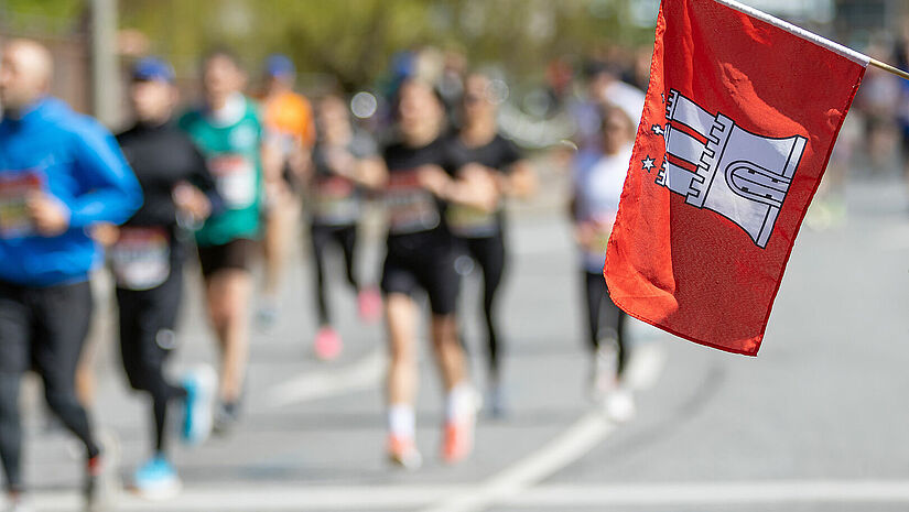 Menschen laufen in Sportkleidung eine Straße entlang. Im Vordergrund schwenkt jemand eine rote Flagge mit dem Hamburg-Wappen.