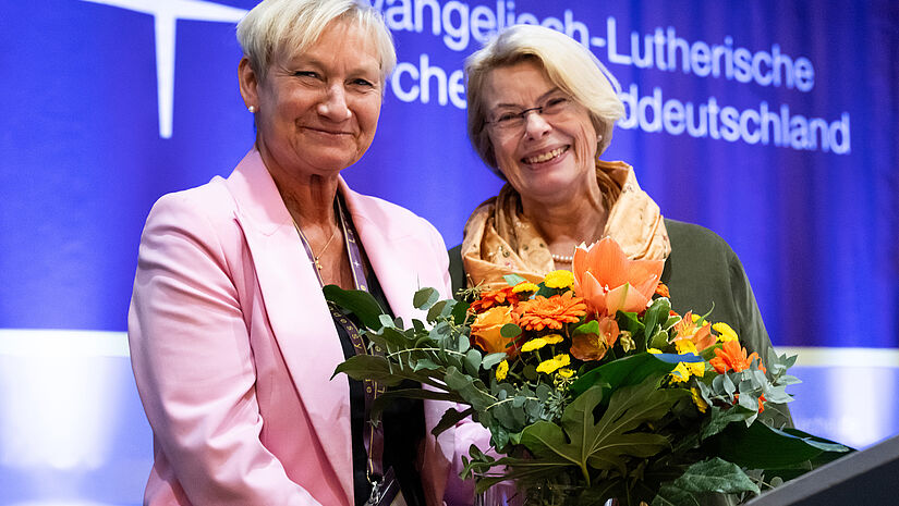 Zwei Frauen lachen in die Kamera. Sie halten einen Blumenstrauß vor sich.