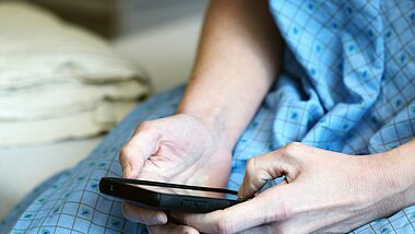 Ein Patient sitzt in einem blauen Nachthemd auf einem Bett, mit seinen Händen tippt er etwas auf sein Smartphone.