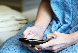 Ein Patient sitzt in einem blauen Nachthemd auf einem Bett, mit seinen Händen tippt er etwas auf sein Smartphone.