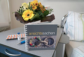 Das Buch im quadratischen Format steht auf einem kleinen Beitisch neben einem Krankenhausbett, im Hintergrund ein Strauß Blumen, ein Kugelschreiber und eine Blisterpackung mit Medikamenten